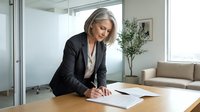 Professional mature Caucasian businesswoman in black suit signing documents at modern office desk with glass walls and contemporary interior design.