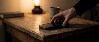 Person with pale skin tone using smartphone on rustic wooden desk at night with warm lamp lighting and coffee cup nearby for work or communication concepts.