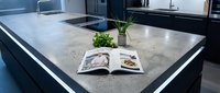 Contemporary kitchen island with concrete countertop, open magazine, potted plant, and modern black cabinetry. Ideal for home design, interior decoration, and lifestyle publications.