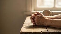 Elderly hands clasped in prayer on wooden table with warm sunlight streaming through window creating peaceful spiritual moment for meditation.