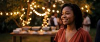 Happy African American woman smiling at outdoor evening party with warm string lights creating bokeh background atmosphere for celebrations and gatherings.