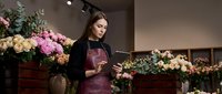 Young Asian female florist in burgundy apron arranging fresh flowers in modern flower shop interior with tablet for online orders and business management.