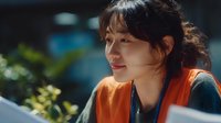 Young Asian woman in orange safety vest smiling outdoors with natural light highlighting her face, conveying confidence and approachability in professional setting.