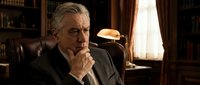 Mature man with gray hair and facial hair, wearing dark business suit, sitting in leather chair in office setting with warm lighting and wooden furniture, contemplative expression with hand near face, suitable for corporate, professional, and business concepts.