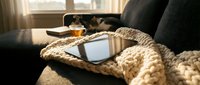 Tablet with blank screen on chunky knit blanket on cozy sofa, warm sunlight, tea and cat in background, hygge home atmosphere.
