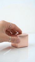 Hand reaching for rose gold metallic cube on white surface with minimalist aesthetic for luxury product design and branding concepts.