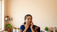 Middle-aged woman of South Asian descent wearing blue traditional attire, smiling thoughtfully with hands near face in home interior with plants and warm lighting.