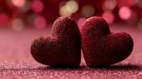Two sparkling red glittery hearts touching on shimmering surface with warm bokeh lights background for Valentine's Day romance celebration.