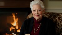 Elegant senior Caucasian woman with white hair sitting in armchair by fireplace, wearing dark cardigan and burgundy top with pearl necklace in cozy home interior.