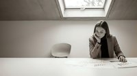 Young professional businesswoman working at modern office desk reviewing documents and charts in minimalist workspace with natural skylight.