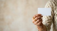 Woman with brown skin holding blank white card, wearing gold rings and beige knit sweater against neutral background for mockup.