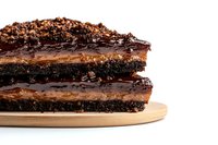 Layered chocolate cake with dark ganache and caramel filling on wooden board, close up view showing rich texture and decadent layers against white background.