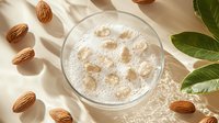 Fresh almond milk in glass with scattered nuts and green leaves on beige background with natural sunlight and soft shadows. Healthy dairy alternative.