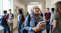 Smiling Muslim female student wearing hijab holding books in busy school hallway with diverse classmates chatting in background during break time.