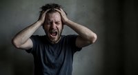 Stressed Caucasian man holding head in hands screaming with emotional breakdown against dark wall background expressing mental health crisis and overwhelming pressure.
