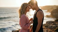 Young couple sharing intimate moment at golden hour sunset by ocean waves on rocky coastline during romantic beach vacation getaway.