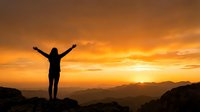 Silhouette of woman with raised arms celebrating success on mountain peak at golden sunset with dramatic orange sky and layered mountain ranges in background.