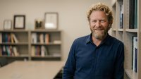 Middle-aged Caucasian man with curly beard smiling in modern office library setting with bookshelves and framed certificates in background.