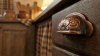 Ornate copper drawer pull with decorative shell design on rustic wooden kitchen cabinet showcasing traditional craftsmanship and vintage hardware details.
