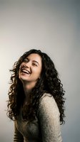 Young Hispanic woman with curly hair laughing joyfully in cozy sweater against neutral background for lifestyle and wellness content.