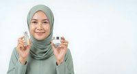 Young Asian Muslim woman in green hijab holding skincare cream jars, smiling at camera on white background for beauty and cosmetic advertising campaigns.