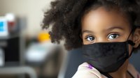 Young African American girl wearing protective black face mask looks at camera with expressive eyes, curly natural hair styled in afro, healthcare and safety concept.