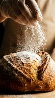 Hands dusting flour over freshly baked artisan bread loaf during food preparation and baking process in kitchen.