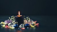 Lit candle surrounded by colorful gemstones and small white flowers on dark background creating romantic atmosphere for meditation and relaxation.