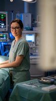 Female surgeon in green scrubs and glasses standing in modern operating room with medical equipment and monitors in background.