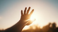 Silhouette of hand reaching toward bright sun in sky, creating sunburst effect through fingers against soft blue backdrop at golden hour.