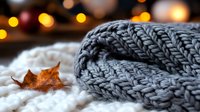 Cozy winter composition with warm gray knitted scarf on soft white blanket, dried autumn leaf and blurred golden bokeh lights creating seasonal atmosphere.
