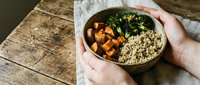 Young woman with light skin tone holding a wooden bowl containing roasted sweet potato cubes, quinoa, and fresh kale on a rustic wooden table, suitable for food blogging, healthy eating content, and nutrition-related designs.
