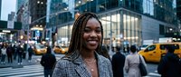 Professional African American businesswoman with braided hair smiling confidently on busy city street with yellow taxis and urban skyscrapers in background.