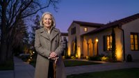 Senior Caucasian woman with gray hair in elegant coat standing confidently outside luxury home at twilight with warm lighting and manicured landscape.