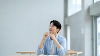 Young Asian man in light blue casual shirt sitting indoors, smiling while looking upward thoughtfully in modern bright office or home space with natural window light.