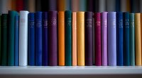 Row of colorful hardcover books arranged on white shelf, spines showing various sizes and thicknesses creating visual rhythm, soft focus background for library setting.