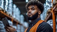 Young African American male warehouse worker in safety vest using digital tablet for inventory management against blurred storage facility background.