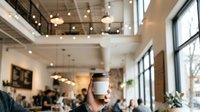 Modern cafe interior with hand holding white coffee cup, blurred customers and bright natural light from windows, suitable for beverage and hospitality marketing.