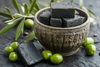 Black charcoal soap bars in rustic ceramic bowl with fresh green olives and olive branches on dark stone surface, natural skincare and beauty concept.