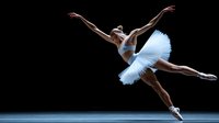 Young African American ballerina in white tutu performing grand jete leap against dark background, showcasing grace and athletic ability in contemporary dance performance.