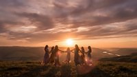 Group of diverse young adults holding hands in circle during golden hour sunset on mountain hilltop with dramatic sky and valley landscape below.