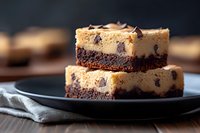 Decadent chocolate chip cookie dough brownies stacked on black plate, featuring rich fudgy brownie base topped with golden cookie dough layer and chocolate chips.