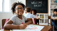 Happy African American elementary school girl with glasses smiling in classroom with welcome back chalkboard message and diverse students studying at desks.
