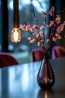 Pink cherry blossoms in dark glass vase with glowing pendant lamp creating cozy atmosphere. Modern interior design with burgundy chairs and moody lighting.