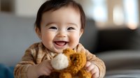 Joyful Asian baby wearing brown sweater laughs while holding cookies, capturing pure childhood happiness and authentic emotional expression in natural home setting.
