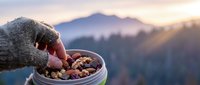 Hand in knitted glove reaching for mixed nuts and dried fruits in metal bowl during mountain sunrise with warm golden light and scenic landscape backdrop.
