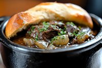Hearty beef stew served in black cast iron pot with tender meat chunks, pearl onions and fresh herbs, topped with crusty artisanal bread for warming comfort food.