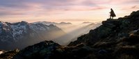 Silhouetted hiker stands on rocky mountain peak at sunrise, overlooking misty alpine valley with snow-capped peaks. Outdoor adventure and wilderness exploration concept.