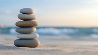 Balanced stack of smooth gray stones on sandy beach at sunset with blurred ocean waves in background, creating zen and peaceful atmosphere.