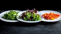 Fresh green kale, purple lettuce leaves, and sliced red bell peppers served on white plates against dark stone background, minimalist food composition.